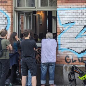 Vier Personen sind auf einem Foto von hinten zu sehen. Sie sind sommerlich bekleidet und stehen vor einem offenen Fenster eines mit Graffiti besprühten Ziegelgebäudes. Sie blicken von außen durch das geöffnete Fenster nach drinnen. Drei sind jünger, darunter eine Person mit langen Haaren. Eine Person hat kurze graue Haare.