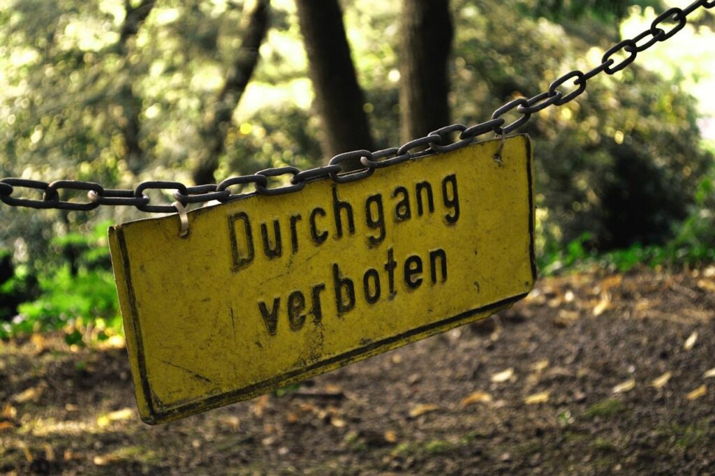 Verbogenes Metallschild an einer Kette, im Hintergrund Waldboden und Bäume. Das Schild enthält schwarze Schrift auf gelbem Hintergrund, Text "Durchgang verboten"